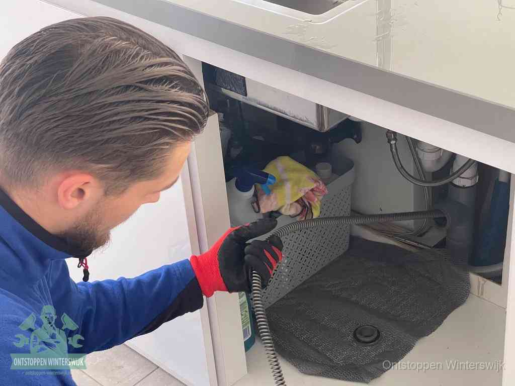 Een monteur voert een flexibele ontstoppingsveer in onder het keukenkastje, langs de sifon, om de gootsteen te ontstoppen in Winterswijk