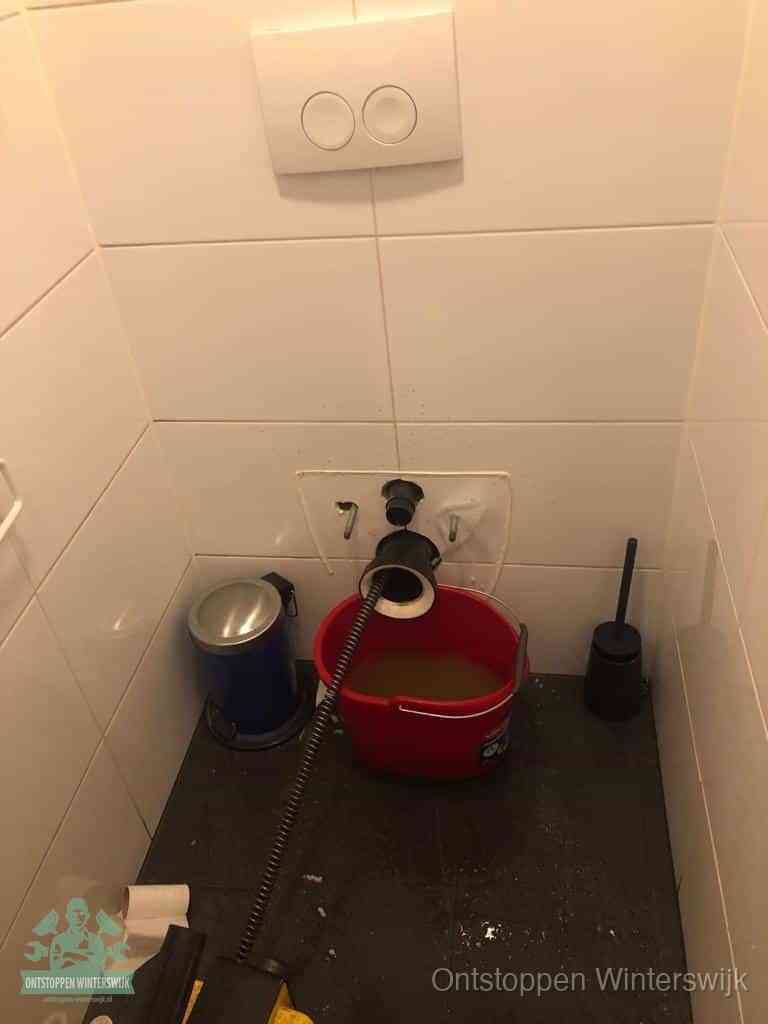 Een ontstopte wand-wc met verwijderde toiletpot waar een ontstoppingsveer in de wanduitgang verdwijnt, het achterhalen waar de verstopping zit in Winterswijk