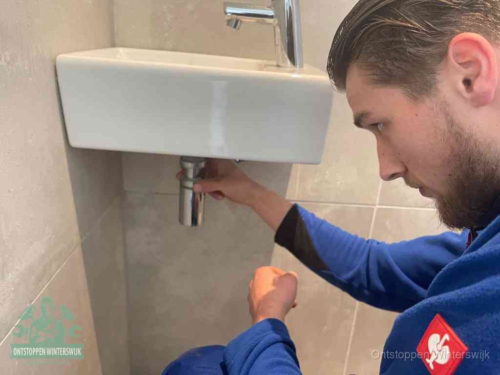 Een monteur draait met de hand de zwanenhalssifon onder een fonteinwasbak in de toiletruimte los om de bron van rioollucht in Winterswijk te vinden